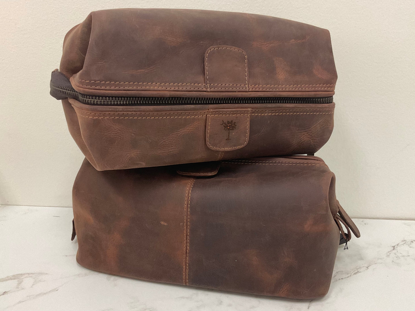 Locally made in Charleston, SC. Two brown leather travel bags stacked on top of each other, one with a zipper and the other with a snap closure.