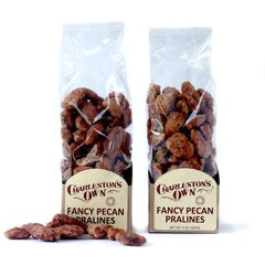Two clear plastic packages of pecan pralines with visible pecan pieces coated in a candy-like texture. The packages have labels with the product name and brand. Locally made in Charleston