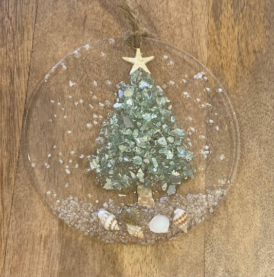 A round, resin and glass Christmas ornament featuring an aqua blue crushed glass Christmas tree with shells and a beach, topped with a gold starfish, on a wooden surface.