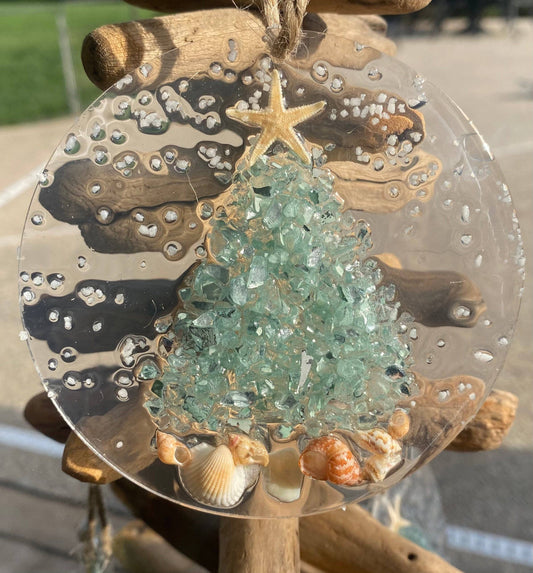 A handmade Christmas ornament featuring an aqua crushed glass tree with seashells and a starfish, set in resin on a clear acrylic round base, with a twine hanger.
