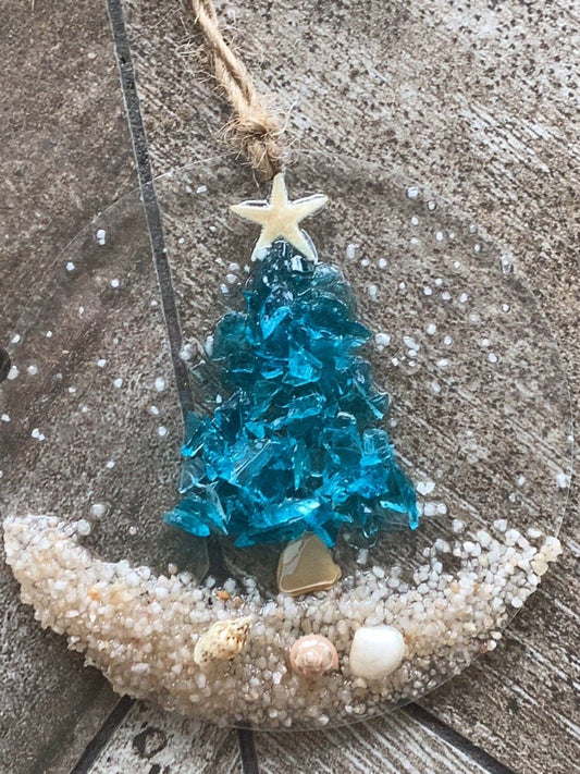A turquoise crushed glass Christmas tree ornament with shells and a starfish on a sandy beach, set in resin and hanging from a twine.